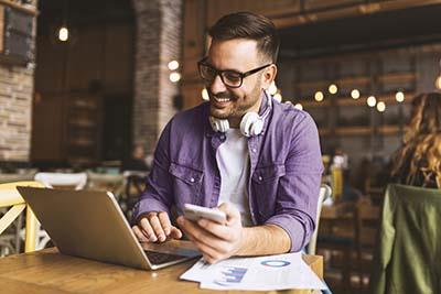 Smiling man using a laptop with a phone in his hand and headphones on his shoulders